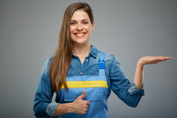 Smiling woman in blue overalls with empty hand for product shows thumb up. Isolated female portrait in builder worker uniform.