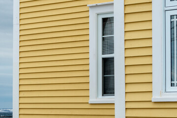 The exterior of a bright yellow narrow wooden horizontal clapboard wall of an old house with vinyl windows. The trim on the glass panes is white in color. The outside boards are textured pine wood.  