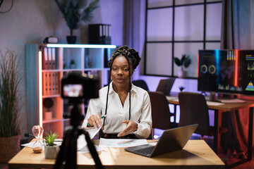 Fototapeta Cheerful confident african american businesswoman, broker in white shirt sitting in front of camera in evening office during recording video for business vlog. obraz
