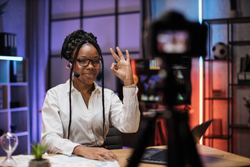 Portrait of attractive confident experienced smart female african american office manager in headset recording video vlog for internet audience in evening office showing sign ok.