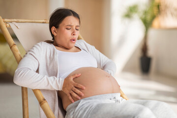 Pregnancy Pain. Pregnant Woman Touching Belly While Sitting In Chair At Home