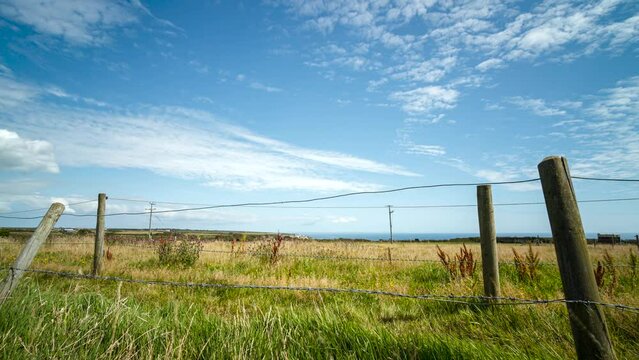 Time lapse shot of a meadow with a sloping fence, some white clouds move through the blue sky.