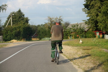 An elderly man rides a bicycle in the village on a hot summer day . Healthy lifestyle and sports in rural areas