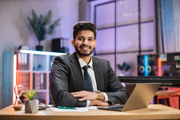 Fototapeta premium Front view portrait of young smiling experienced smart stylish bearded hindu office manager in suit sitting at table using laptop in evening office.