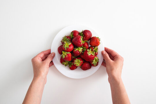 Female hands hold a plate with strawberries on a white table background. Top view, flat lay