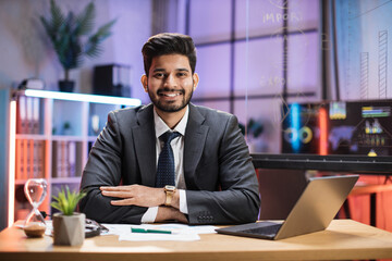 Fototapeta na wymiar Close up portrait of smiling confident indian financial expert office worker sitting at table in formal suit working with laptop in evening modern office.