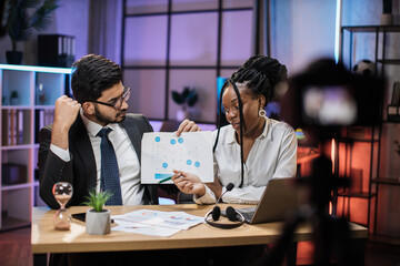 Two multicultural company workers arab financial expert and african woman office worker using laptop, showing paper report with graphs and charts during video meeting with colleagues in office.
