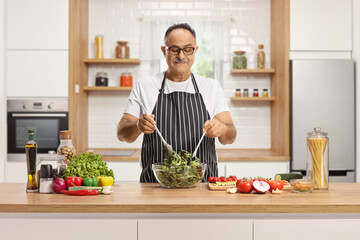Man wearing an apron behind a kitchen counter mixing a salad