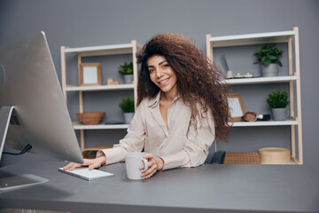 Cheerful smiling tanned adorable curly Latin businesswoman in linen shirt welcomes new colleagues. Copy space. Attractive freelancer work from home office using modern desktop computer