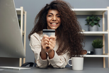 Overjoyed tanned adorable curly Latin woman in linen shirt holding her favorite credit card in home office interior. Copy space Mockup Banner. Shopaholic use modern smartphone for online shopping