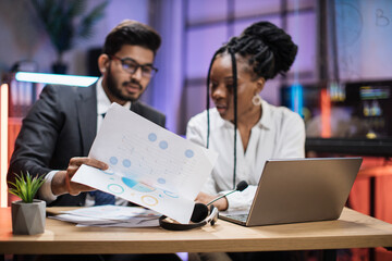 Focus on papers with report of two multicultural company workers arab expert and african woman office worker using laptop, showing graphs and charts during video meeting with colleagues in office.