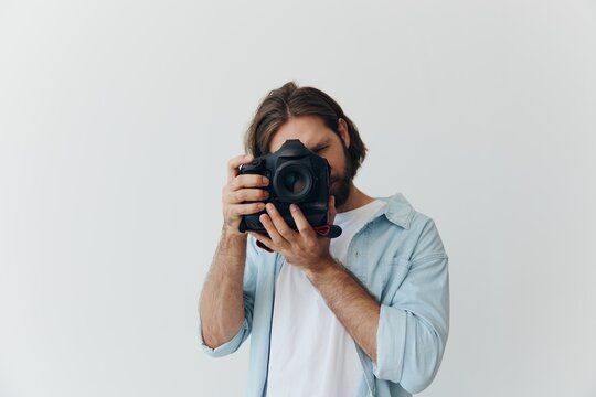 A male hipster photographer in a studio against a white background looks through the camera viewfinder and shoots shots with natural light from the window. Lifestyle work as a freelance photographer