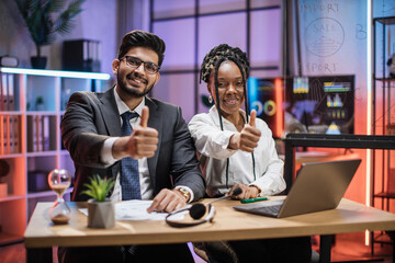 Two multi ethnic economists, young smiling experienced african american female and arab male office manager in formal wear sitting at table using laptop in evening office showing thumbs up.