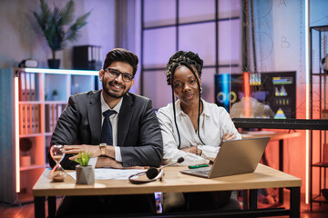 Two multi ethnic economists, young smiling experienced african american female and arab male office manager in formal wear sitting at table using laptop in evening office.