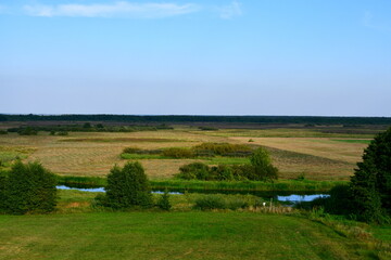 A view from the top of a tall hill with numerous trees, shrubs, and forest formations visible next to some fields, meadows, and pasturelands spotted on Polish countryside in summer