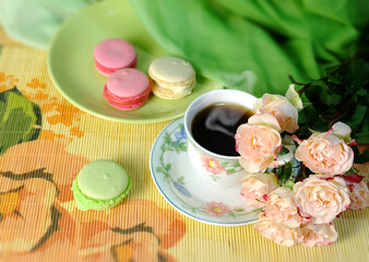 A cup of coffee, macaroons, and a bouquet of pink roses on a green chiffon background, a horizontal holiday composition with sweets for a greeting card, poster, etc.