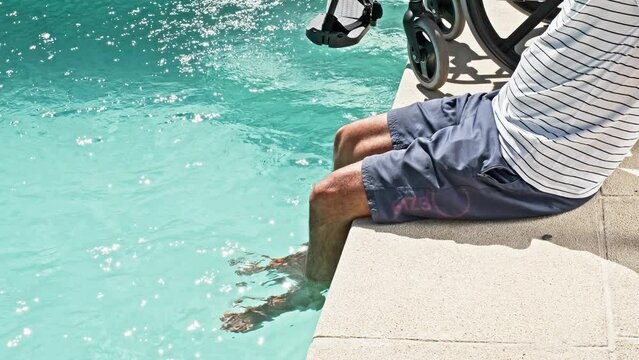 person with a disability sitting next to his wheelchair with his feet in the pool sticks his legs out of the water