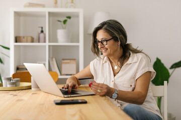 Smiling female shopping online via laptop at home - obrazy, fototapety, plakaty