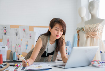 Happy Asian woman dressmaker or designer seamstress tailor with a tape measure using laptop and sketches design for trendy fashion in the studio office.