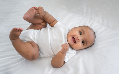 Portrait of happy African American mixed race cute girl baby lying down on white bed and look on camera in a bedroom at home.