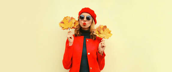 Autumn portrait of beautiful young woman model with yellow maple leaves blowing her lips with lipstick wearing red french beret on gray background