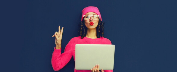 Portrait of stylish modern young woman working with laptop wearing pink colorful clothes on dark blue background, banner blank copy space for advertising text