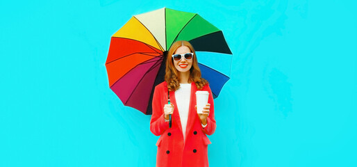Autumn portrait of young woman with colorful umbrella drinking juice or cup of coffee wearing red jacket on blue background