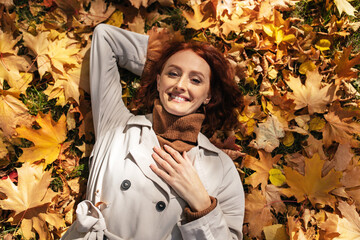 Glad happy caucasian millennial red-haired female in raincoat lies on ground in park with yellow leaves - obrazy, fototapety, plakaty