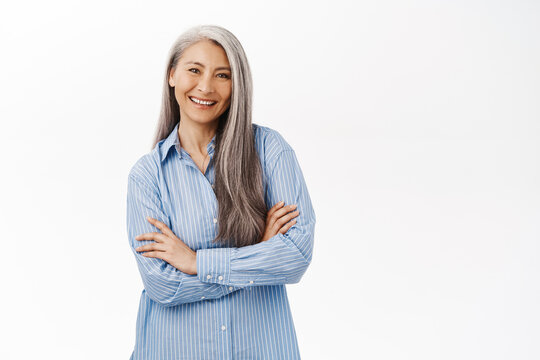 Smiling asian woman, senior lady looking with confidence, standing in power pose against white studio background