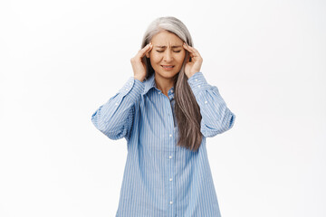 Portrait of japanese senior woman having headache, touching head and grimacing from pain, migraine, standing over white background