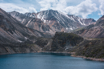 Obraz premium Deep mountain lake of phantom blue color among sharp rocks and high mountains in changeable weather. Wonderful dramatic view to deep blue mountain lake and large sunlit rocky cliff under cloudy sky.
