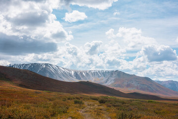 Motley autumn landscape with hills on high plateau and sunlit snowy mountain range under dramatic cloudy sky. Vivid autumn colors in mountains. Sunlight and shadows of clouds in changeable weather.