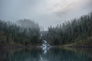 Mountain creek flows from forest hills into glacial lake in mysterious fog. Small river and coniferous trees reflected in calm alpine lake in early morning. Tranquil misty scenery with mountain lake.