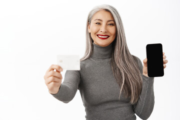 Bank and people concept. Happy asian lady showing mobile phone screen, credit card, demonstrates app interface on smartphone, standing over white background