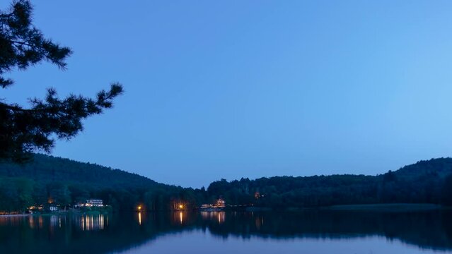 Time lapse shot from day to night of a small lake in the woods of France. The clear sky shows the stars. Some lights appear at the waterfront.