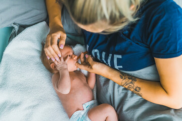 Caucasian woman with blond hair lying in bed with her newborn baby boy caressing his head and letting him suck her thumb. Caring for infant. Motherhood. Horizontal indoor shot. High quality photo