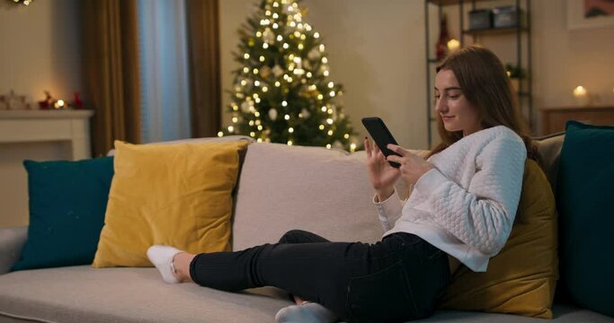 Winter evening. After work, the girl sits on couch in the living room. The beauty holds phone in hands, looks at funny photos and smiles. The room is cozy, shimmering Christmas tree.