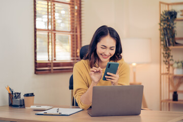 Attractive brunette Asian woman using mobile phone, smile and happy while sitting at working desk in modern office.