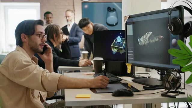 A 3D model artist discusses a project over the phone at a game and app development company. In the office, other employees work at computers.