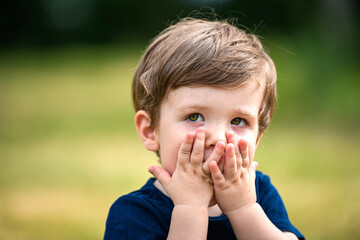 A beautiful little Caucasian boy, looking scared or guilty, cover his face with his hands, hiding,...