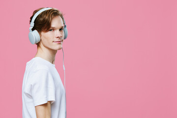 portrait of a cute red-haired guy in a t-shirt listening to music in bright headphones smiling looking at the camera. Studio photo on a solid pink background with empty space for an advertising insert