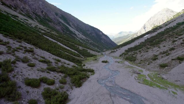 Aerial drone footage flying along a river through a dramatic glacial valley surrounded by a steep mountains and pine trees with patches of snow in Switzerland.