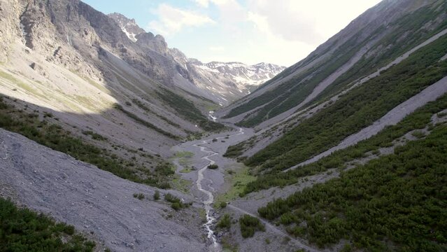Aerial drone footage flying down and through a dramatic glacial valley surrounded by a steep mountains and pine trees with patches of snow and an alpine river in Switzerland.
