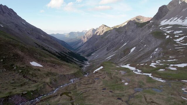 Aerial drone footage facing down a glacial valley and slowly reverseing through a dramatic, jagged mountain landscape with residual patches of snow and alpine meadows in Switzerland.