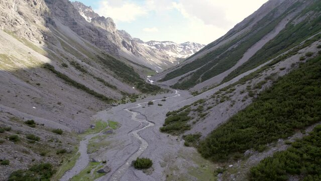 Aerial drone footage slowly descending into a dramatic glacial valley surrounded by a steep mountains and pine trees with patches of snow and an alpine river in Switzerland.