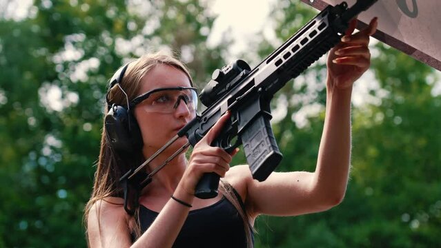Caucasian woman wearing safety headphones and goggles looking at camera holding submachine gun. Outdoor shooting range. Horizontal shot. High quality 4k footage