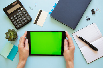 Tablet computer with green screen and female hands on blue background. Top view of desktop with various office supplies. Flat lay, mock up.