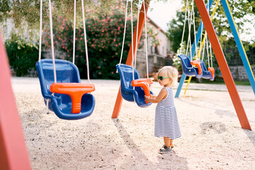 Little girl stands near the swing holding on the seat. Side view. High quality photo