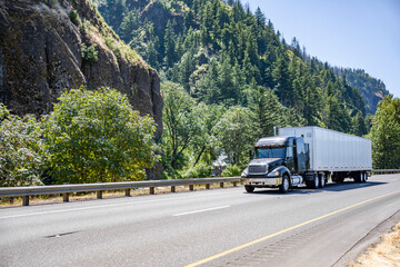 Old popular black big rig semi truck transporting cargo in dry van semi trailer running on the wide highway road