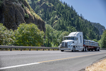 White bonnet big rig semi truck transporting fastened pipes on the flat bed semi trailer driving on the road with rocks and forest on the side